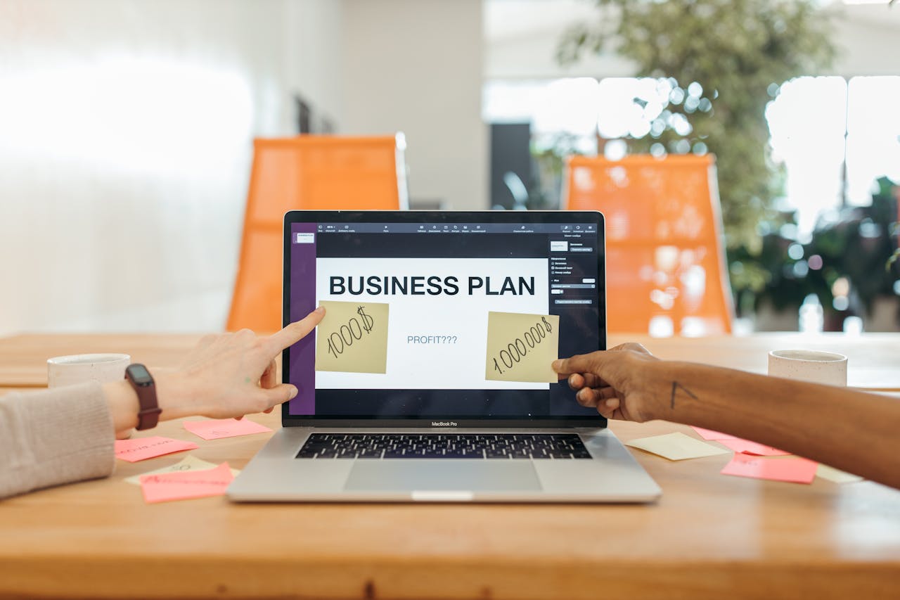 Home Two professionals strategizing a business plan on a laptop with sticky notes and profit goals.